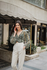 Fototapeta premium stylish young woman with brunette long hair and makeup smiling while talking on smartphone and standing in trendy outfit with handbag on chain strap near blurred fancy restaurant in Istanbul