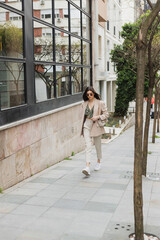 young brunette woman in stylish sunglasses and trendy outfit with white pants and beige blazer walking with handbag near trees and modern building on street in Istanbul