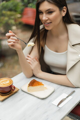 smiling woman with long hair holding fork with cheesecake next to cup of cappuccino with coffee art on bistro table while sitting inside of modern cafe in Istanbul