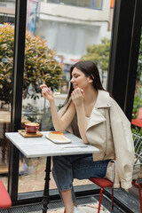 happy young woman with long hair eating cheesecake near cup of cappuccino with coffee art on bistro table while sitting in leather jacket next to window inside of modern cafe in Istanbul