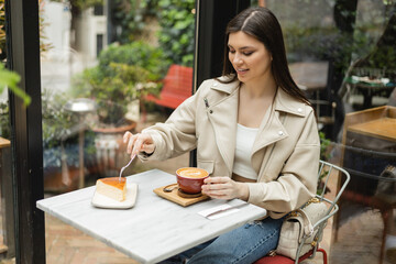 happy woman with long hair sitting in leather jacket next to window and bistro table while holding cup of cappuccino and fork above cheesecake inside of modern cafe in Istanbul