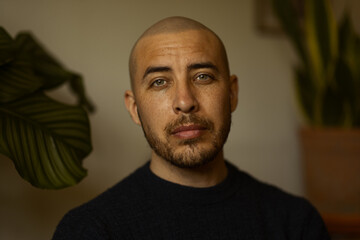 Obraz premium Intense portrait of a handsome bald man with a beard and striking green eyes, looking directly at the camera with a calm expression against a soft indoor background with green plants and shadows.