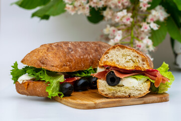 Sandwich from baked bread, mozzarella cheese, salami, prosciutto, olives, sauce, salad leaves, on a wooden bottom