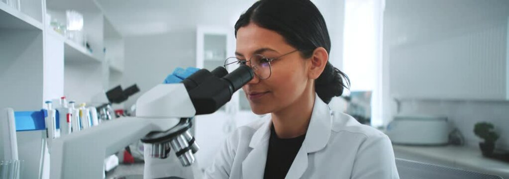 A young scientist woman with glasses looking under the microscope writing in a notebook in the laboratory. Medicine, biotechnology research. Pharmacology