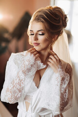 Fototapeta premium A blonde bride in a long, lacy petticoat is standing in her room, posing next to her wedding dress. Beautiful hair and make-up, open bust