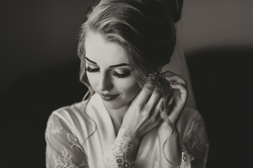 Black and white photo. A blonde bride in a long, lacy petticoat is standing in her room, posing by the mirror, putting on her earrings. Beautiful hair and makeup, open bust. Wedding portrait.  