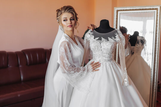 A blonde bride in a long lace robe and white shoes stands in her room next to her wedding dress, posing by the mirror. Beautiful hair and make-up, open bust. Wedding portrait. Open legs