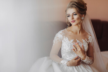 Blonde bride in long sleeve lace dress standing in her room, posing and putting on her engagement ring. Beautiful hair and makeup, open shoulders. Wedding portrait.