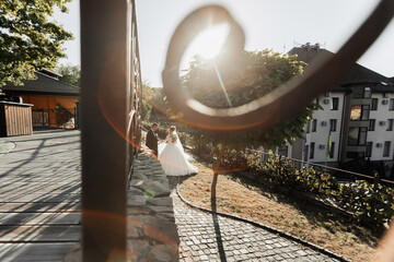 Wedding portrait. The groom in a black suit and the blonde bride are walking holding hands near a stone wall under a tree. A white, long dress in the air. Sun rays in the photo