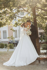 Obraz na płótnie Canvas Wedding portrait. A groom in a black suit and a blonde bride are standing, hugging and kissing under a tree. Photo session in nature. A beautiful ray of the sun in the photo. Beautiful hair and makeup