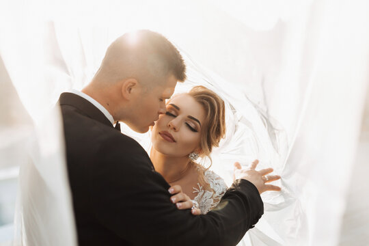 Wedding portrait. The groom in a black suit and the blonde bride are hugging, wrapped in the bride's veil. Long dress in the air. Photo session in nature. Beautiful hair and makeup