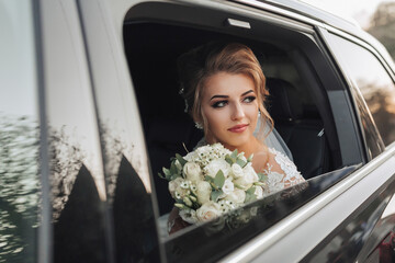A blonde bride, smiling sincerely, sits in a black car on her wedding day with a bouquet of white...