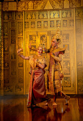 Obraz premium Vertical image of beautiful Asian woman wear Thai traditional dress action of dancing together with man stand in front of Thai painting on public place wall.