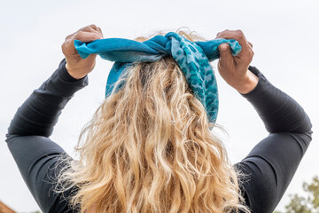 Blonde woman from behind, ties her hair with a green and blue scarf
