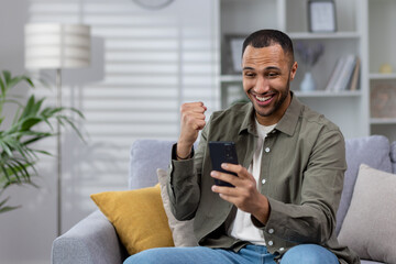 Young smiling Muslim man using phone while sitting at home on sofa, playing online games, placing bets, enjoying money win, celebrating, showing victory gesture with hand