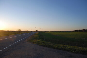 Asphalt road at sunset. Spring evening in a clear sky, the sun leaned low to the horizon, a gray asphalt road passes among the green fields, trees are visible in the distance. no clouds in the sky.