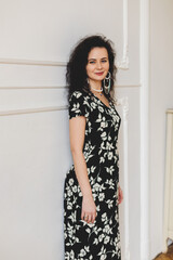 Fototapeta premium Portrait of young beautiful caucasian brunette curly woman in gorgeous flower black and white print dress standing near wall. Elegant lady. Girl look thoughtful and dreamy. Happy smiling woman.