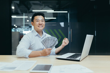 Fototapeta na wymiar Portrait of a happy Asian man working in the office on a laptop, holding a fan of cash, dollar bills in his hand. shows a victory gesture with his hand to the camera, celebrates.