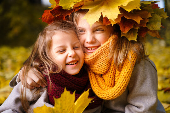 Beautiful little girls in bright yellow red orange autumn warm knitted scarf snood with head wreath of maple leaves,foliage walking in forest. Fun playing in park. Outdoor family activity in nature