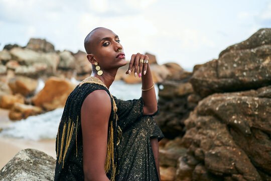 Non-binary black person in luxury dress on rocks in ocean. Trans ethnic fashion model wearing jewellery dressed in posh gown poses gracefully in tropical seaside location portrait. Pride month.