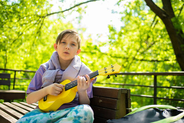 a boy plays a small guitar, a yellow ukulele on a bench in the park