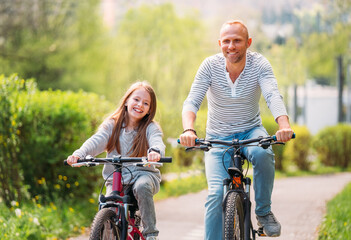 Obraz premium Portraits Smiling father with daughter during summer outdoor bicycle riding. They enjoy togetherness in the summer city park. Happy parenthood and childhood or active sport life concept image.