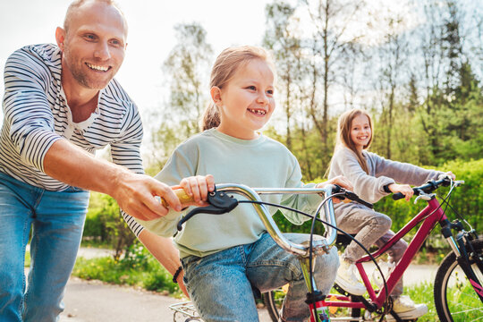 Smiling father with two daughters during outdoor walk. He teaching younger girl to ride a bicycle. They enjoy togetherness in the summer city park. Happy childhood concept image..