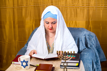 A Jewish woman in a kisuy rosh headdress and a prayer veil reads a siddur prayer in the synagogue.
