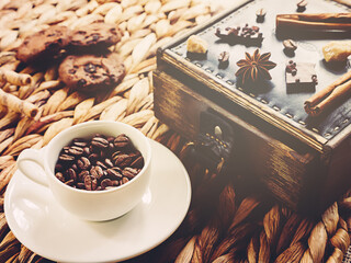 White cup with coffee beans and chocolate chip cookies on a wicker background. on wooden. box of pieces of sugar, chocolate, sweet sticks and cinnamon. selective focus