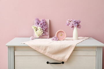 Vase of beautiful fragrant lilac flowers with frame and clock on commode near pink wall