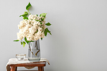 Vase with lilac flowers and candle on stool near light wall