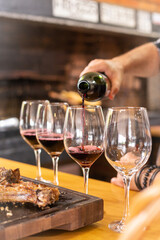 A white man hand serving a red wine bottle in four wine glasses in a row next to some gaucho traditional meat on a rustic wooden board in a familiar home or restaurant for lunch or dinner event