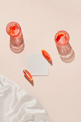 Two glasses of fresh lemonade, water or cocktail, slices of red blood orange with white card note on pastel beige backgound with silk cloth and shadow. Summer refreshment concept. Sunlit flat lay. 
