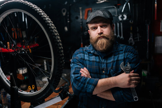 Portrait of bearded cycling mechanic male holding wrench in hand standing by bicycle in repair workshop with dark interior, looking at camera with serious expression. Concept of bicycle maintenance.