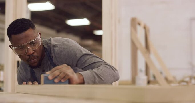 Carpentry, design and focus with a black man sanding a plank in a factory for professional woodwork. Safety goggles, manual labor and expertise with a serious male carpenter working in his workshop