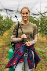 Obraz premium A pretty young woman holding a sprayer for plant protection stands in a fruit farm.