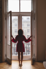 Young woman standing on the balcony and looking at the city. Woman wear short kimono, summer kimono, cotton warp dress, red robe. Curly hair girl. Sexy minimalistic outfit at home.