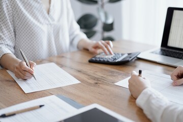 Woman accountant using a calculator and laptop computer while counting taxes with colleague at wooden desk in office. Teamwork in business audit and finance.