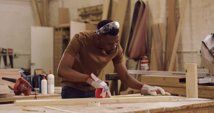 Carpentry, design and a man measuring a plank of timber in his factory for professional woodwork. Safety goggles, manual labor and expertise with a male carpenter using a tape measure in his workshop