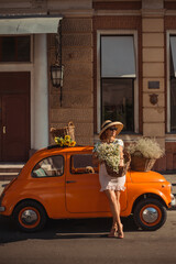 Young beautiful woman in a hat and sunglasses holding a basket of flowers, leaning on a vintage car