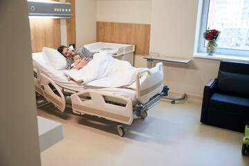 Young mom resting with her sick infant in pediatric ward