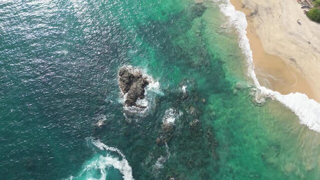 Vista a&eacute;rea del mar de color turquesa y verde y la playa con arena de color beige. Se ven las rocas de color caf&eacute; y la espuma de las olas rompiendo en ellas de color blanco