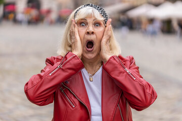 Oh my God, Wow. Senior woman looking surprised at camera with big open eyes, shocked by sudden victory, game winning, lottery goal achievement, good news outdoors. Elderly grandmother in city street