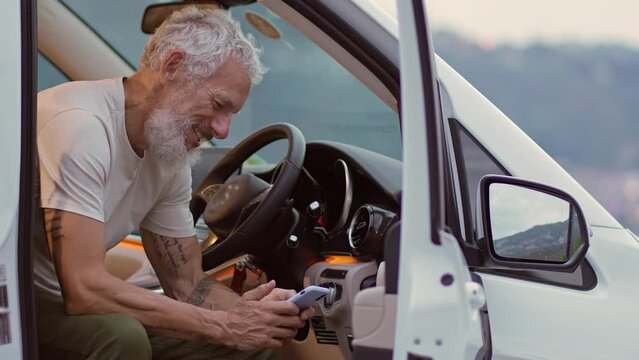 Happy older man sitting in rv camper van using mobile phone. Smiling mature active traveler holding smartphone texting enjoying view and wireless internet in camping, booking campervan.