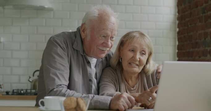 Elderly couple man and woman talking and looking at laptop screen discussing content in kitchen at home. Modern technology and retirement concept.