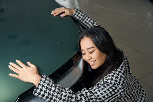 Exalted young asian woman hugging shining car hood