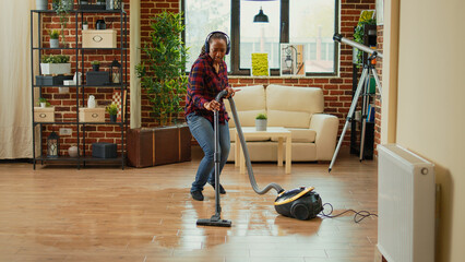 African american housewife using vacuum cleaner and listening to music on headset, enjoying modern...