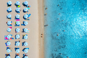 Aerial view of colorful umbrellas on sandy beach, swimming people in blue sea at summer sunny day. Sardinia, Italy. Tropical seascape. Travel and vacation. Top drone view of ocean with azure water