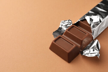 Delicious chocolate bar wrapped in foil on light brown background, closeup. Space for text