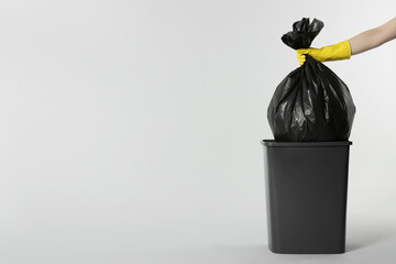 Janitor in rubber glove holding trash bag full of garbage over bucket on light grey background,...